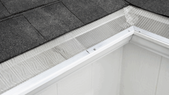 Close-up view of a house roof corner featuring dark asphalt shingles and a white rain gutter with Gutterglove mesh guard, utilizing V-Bend Technology to prevent debris from entering the gutter.